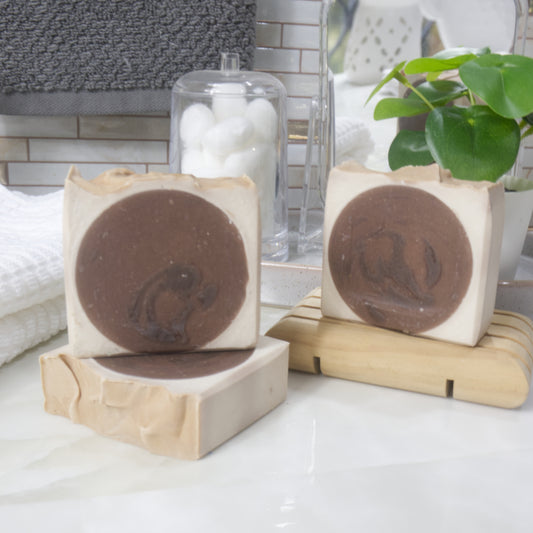 A collection of 3 soaps are displayed on a marble bathroom counter along with a towel, plant and jar of cotton balls.  One soap lays flat on the bench with another standing upright on it.  To the back left is a third soap standing on a wooden soap dish.  The soaps have a dark brown circle in the center with a cream surround of soap