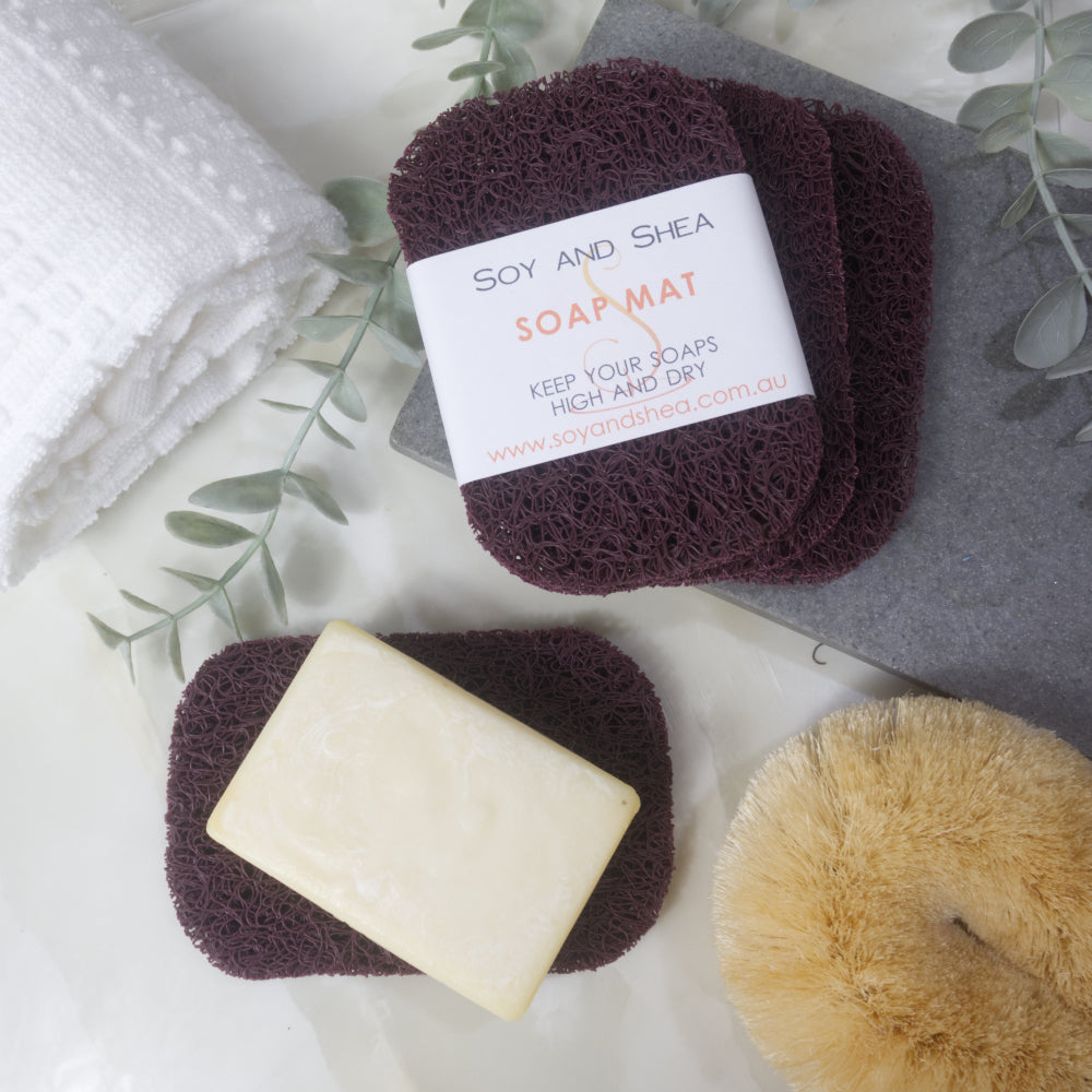 A flatlay image showing a collection of Maroon Soap mats, displayed on a bathroom countertop along with a white towel, body brush and greenery.  Three soap mats are stacked upon each other in a fan shape on a grey stone tray with the top one featuring a product label.  A fourth mat lies to the bottom of the image with a cream soap lying on it.