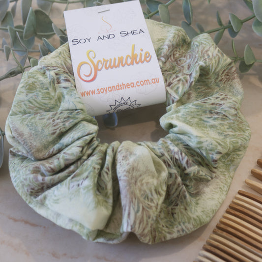  A colourful scrunchie with a cardboard tag lies on a stone counter propped against a plant with the edge of a wooden comb showing.  The scrunchie has a water colour image of thick grass growing in a meadow.