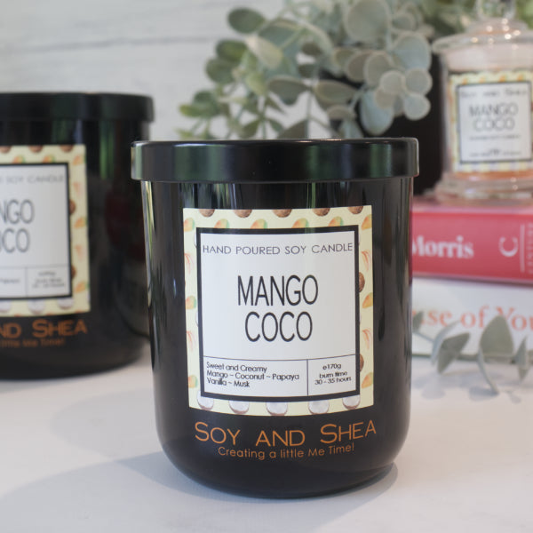 A black candle jar with a lid embossed with the Soy and Shea Logomark sits on a white counter with a label reading MANGO COCO and 170g, sits on white counter with a stack of candles in the background