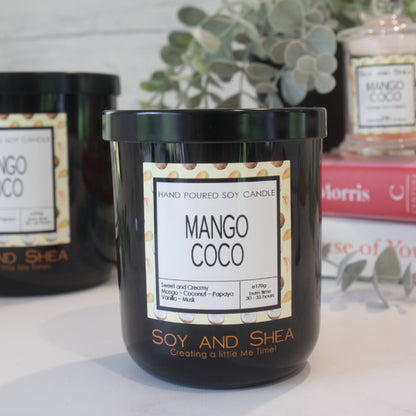 A black candle jar with a lid embossed with the Soy and Shea Logomark sits on a white counter with a label reading MANGO COCO and 170g, sits on white counter with a stack of candles in the background