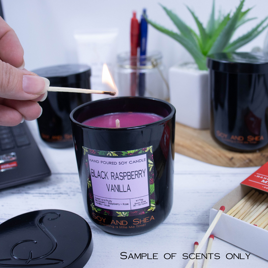 A hand with a lit match reaches in to light a candle in a black jar with purple wax.  It is on an office desk surrounded by matches, stationery, laptop and greenery.