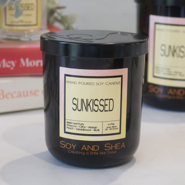 A black candle jar with a lid embossed with the Soy and Shea Logomark sits on a white counter with a label reading SUNKISSED and 170g, sits on white counter with a stack of candles in the background