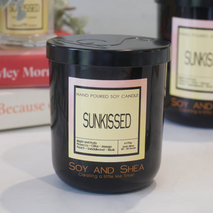 A black candle jar with a lid embossed with the Soy and Shea Logomark sits on a white counter with a label reading SUNKISSED and 170g, sits on white counter with a stack of candles in the background