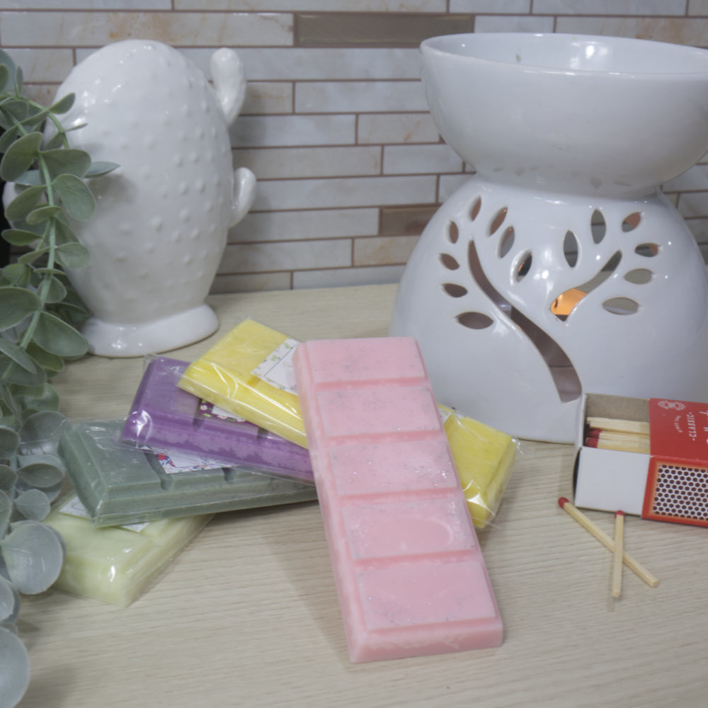 A selection of packaged wax melts with a pink on foremost, next to a  white ceramic diffuser, and matches on a wooden surface.