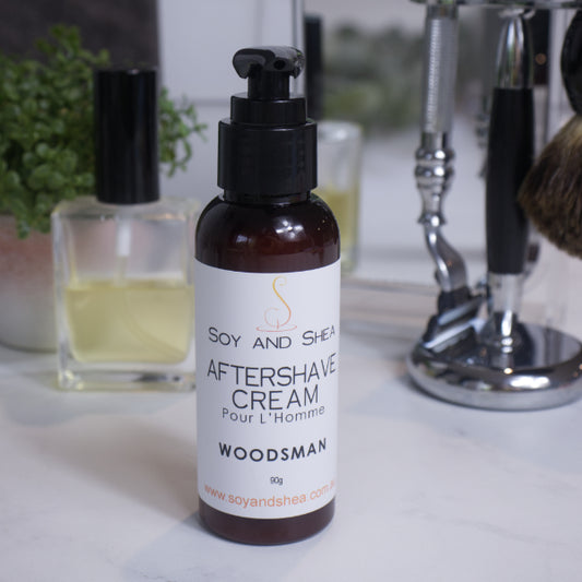 A bottle of Woodsman Aftershave Cream by Soy and Sheen, with a pump dispenser, placed on a bathroom shelf alongside shaving brushes and other items.