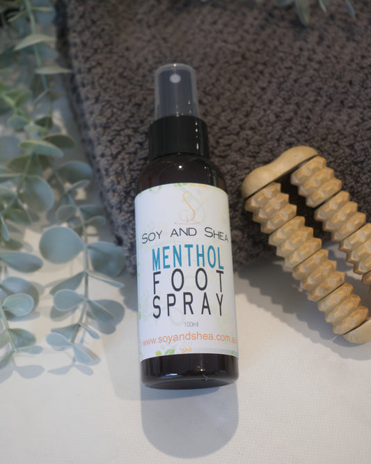 An amber bottle with atomiser lidand a white label reading MENTHOL FOOT SPRAY rests agains a grey towel next to a foot massage roller and green, vine like plant