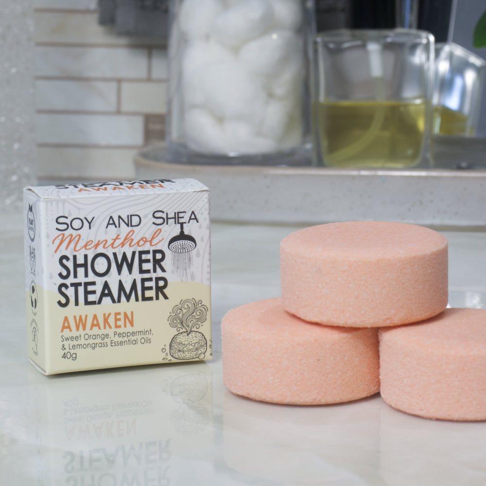 A box of Soy and Shea Menthol Shower Steamer labeled 'Awaken' sits on a countertop next to three round, pale orange shower steamer discs.