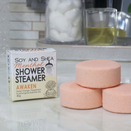 A box of Soy and Shea Menthol Shower Steamer labeled 'Awaken' sits on a countertop next to three round, pale orange shower steamer discs.