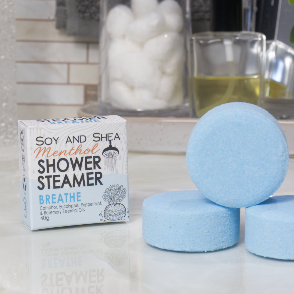 A box of Soy and Shea Menthol Shower Steamer labeled 'Breathe' sits on a countertop next to three round, blue shower steamer discs.