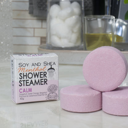 A box of Soy and Shea Menthol Shower Steamer labeled 'Calm' sits on a countertop next to three round, pale mauve shower steamer discs.