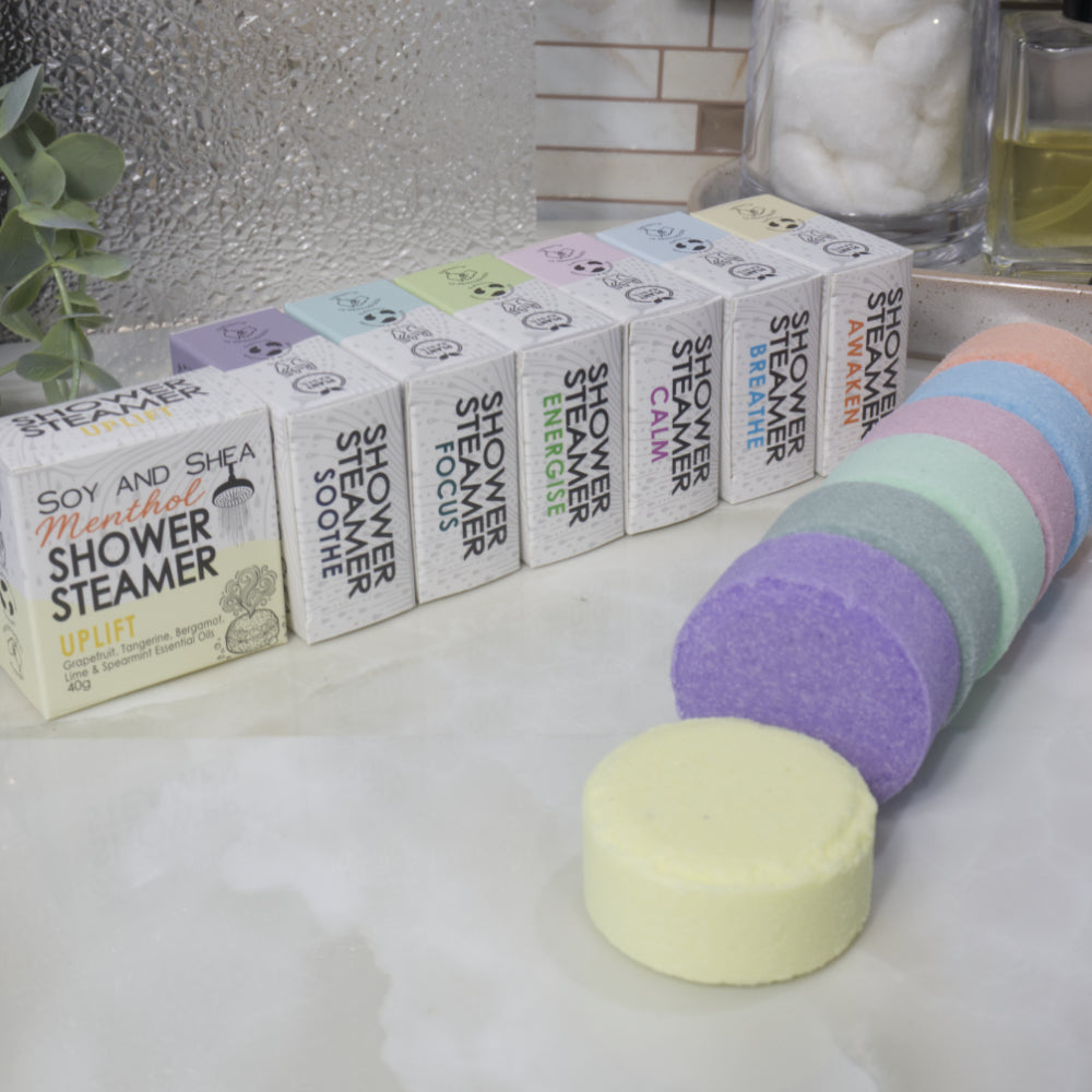 A set of Soy and Shea Menthol Shower Steamer boxes, showing multiple white boxes with colorful labels lined up next to a stack of round pastel Shower Steamers in purple, mint, pink, peach, and pale yellow on a white marble surface.