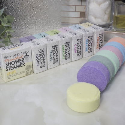 A set of Soy and Shea Menthol Shower Steamer boxes, showing multiple white boxes with colorful labels lined up next to a stack of round pastel Shower Steamers in purple, mint, pink, peach, and pale yellow on a white marble surface.