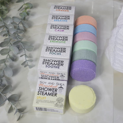 Seven boxed shower steamer variants are arranged next to seven round shower steamers in different pastel colors on a light marble surface, with decorative eucalyptus leaves and a white towel nearby.