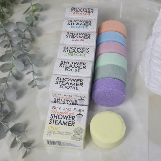 Seven boxed shower steamer variants are arranged next to seven round shower steamers in different pastel colors on a light marble surface, with decorative eucalyptus leaves and a white towel nearby.