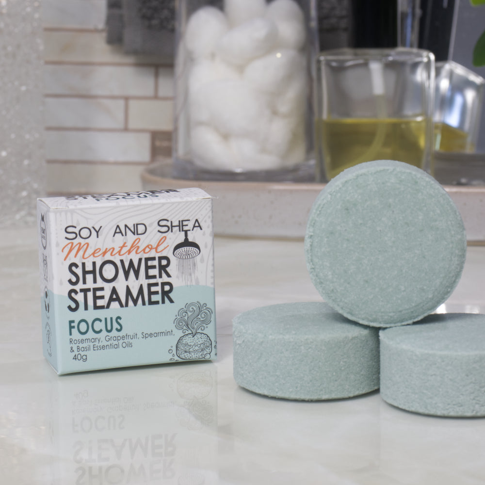 A box of Soy and Shea Menthol Shower Steamer labeled 'Focus' sits on a countertop next to three round, pale dark green shower steamer discs.