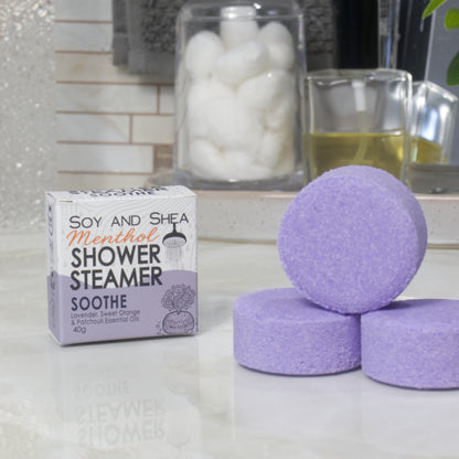 A box of Soy and Shea Menthol Shower Steamer labeled 'Soothe' sits on a countertop next to three round, purple shower steamer discs.