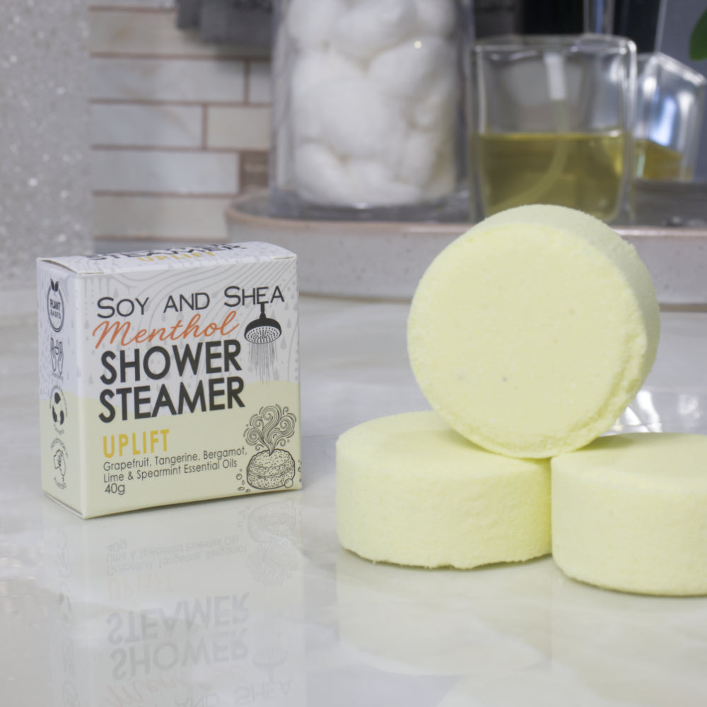 A box of Soy and Shea Menthol Shower Steamer labeled 'Uplift' sits on a countertop next to three round, pale yellow shower steamer discs.