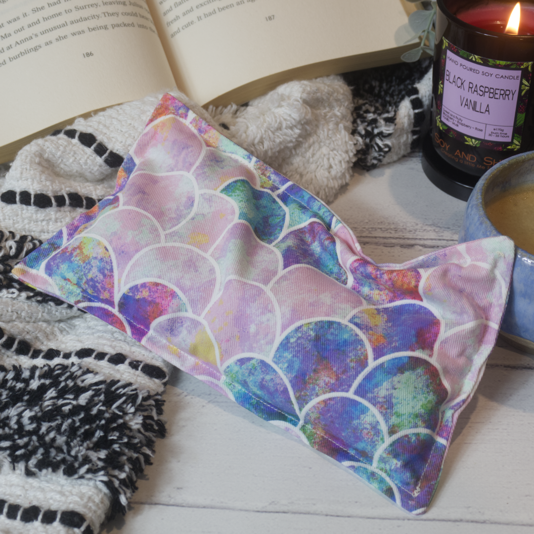 A mini rectangular heat pack rests against a black and white blanket and open book.  Next to it is a burning candle and filled coffee cup evoking a comfy scene.   The fabric features of design of mermaid scales in shades of pink and purple