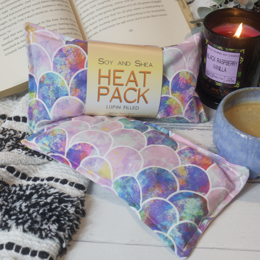 Two Mini Heat packs are stacked upon each other, one with a belly band label reading HEAT PACK LUPIN FIlled.   These are resting on a black and white blanket with open book, candle and coffee cup evoking a cosy feel.    The Heat packs are in light shades of pink and purple covered with a mermaid scale pattern