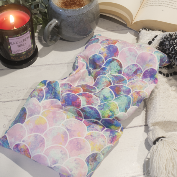 A small rectanglular Heat Pack rests on a wooden table top along with a black and white blanket, open book, burning candle and filled coffee cup evoking a cosy scene.  The Heat pack has a multicolored mermaid tail design in pastel shades of pink and purple