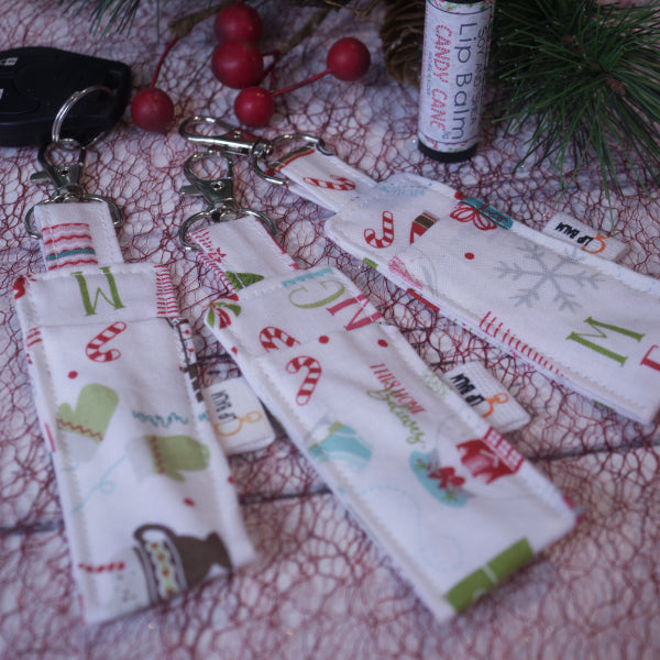 A selection of three clip balms showing variations in fabric design, lie on a white surface with a red mesh with one Clip Balm attached to a car key with a lobster clip.  A lip balm stands near-by along with some Christmas greenery.  The Clip balms are white with various winter and christmas elements including candy canes, mittens, snowglobes and hot drinks