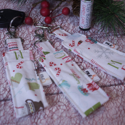 A selection of three clip balms showing variations in fabric design, lie on a white surface with a red mesh with one Clip Balm attached to a car key with a lobster clip.  A lip balm stands near-by along with some Christmas greenery.  The Clip balms are white with various winter and christmas elements including candy canes, mittens, snowglobes and hot drinks