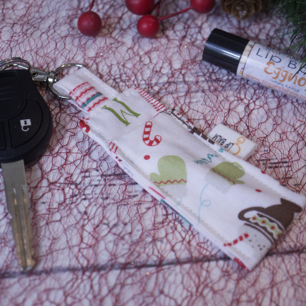 A lip balm holder lies on a white surface covered in red mesh and is attached to a car key with a "lobster claw" clip.  To the side is a tube of lip balm surrounded by christmas greenery and red berries.  The Clip Balm is white with various winter themed images including mittens, hot drink and candy cane