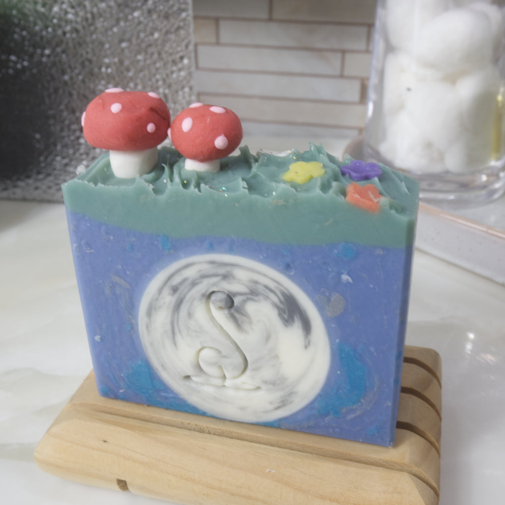 A soap stands on a wooden soap rack on a bathroom counter which has perfume bottle, plant and jar of cotton.   The soap features a bright blue main colour with a white and silver circle in the middle made to look like a full moon.  There is a green layer across the top of the soap which as 3D soap embellishments of two red mushrooms and some flowers