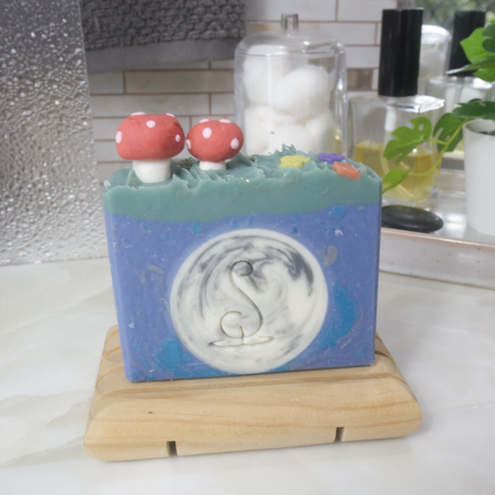 A soap stands on a wooden soap rack on a bathroom counter which has perfume bottle, plant and jar of cotton.   The soap features a bright blue main colour with a white and silver circle in the middle made to look like a full moon.  There is a green layer across the top of the soap which as 3D soap embellishments of two red mushrooms and some flowers