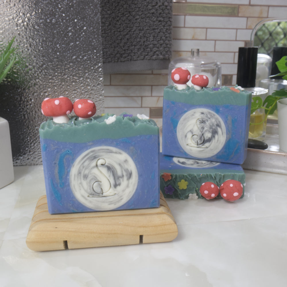 A selection of three soaps displayed on a bathroom bendch along with a bottle of perfume, plant and jar of cotton balls.  One soap sits on a wooden rack to the front left of the image, another two are displayed behind with one laying flat showing the top of the soap with a third standing on top.  Each soap features a round white moon with grey streaks in the center surrounds by a bright blue soap.  The top has a layer of green soap and is finished with a large and small red mushroom and small flowers