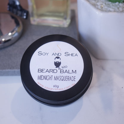 A close-up of a black tin of Soy and Shea's Midnight Masquerade Beard & Mo Balm resting against a stone tray on a marble bench surrounded by blurred bathroom accessories.
