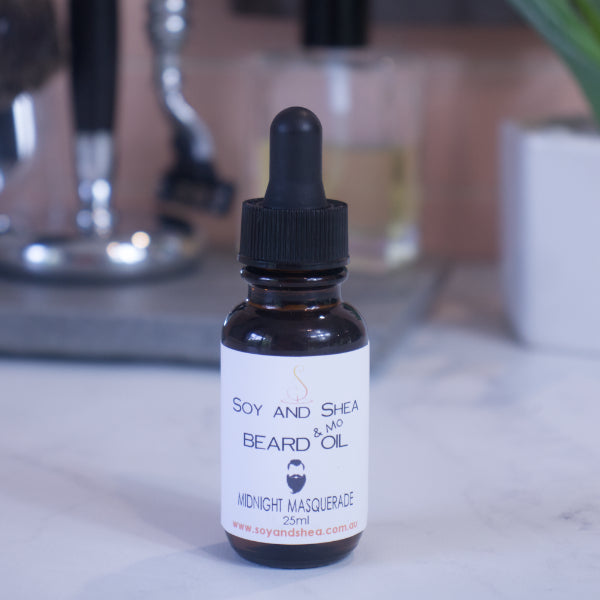 A small brown bottle with a dropper lid, labeled "Soy and Shea Midnight Masquerade Beard & Mo Oil," sits on the marble surface with shaving accessories on a blurred background
