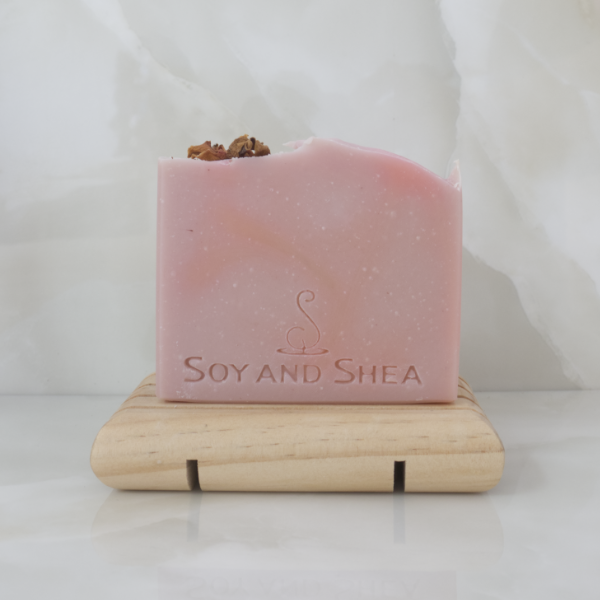 A dusky pink soap bar with a small amount of dried rose petals on the top left corner, sits on wooden soap dish on a marble tile background