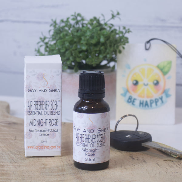 A small amber bottle with black lid labelled AIR FRESHENER SCENT stands on a wooden surface next to the box packaging.  In the background is a car key, air freshener and greenery.