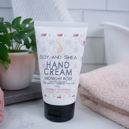 A tube of Soy and Shea hand cream with a floral design, labeled 'Midnight Rose', placed on a surface next to a plant and a towel.  The tube is white with a black flip-top lid.
