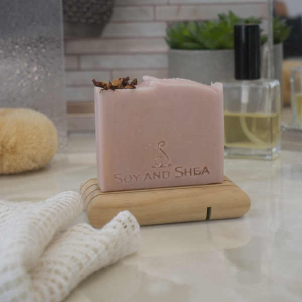 A dusky pink bar of soap with dried rose petals on the top left sits on a wooden soap dish with a bottle of perfume and a fluffy towel in the background.