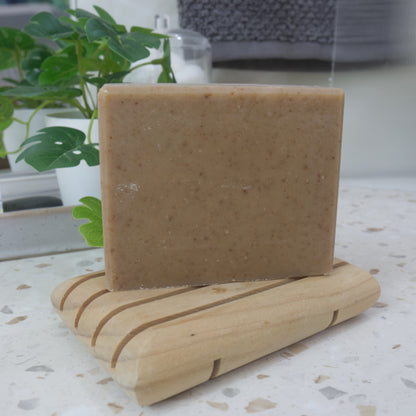 A tan bar of soap with brown flecks stands upright on a wooden soap rack.  It is on a bathroom countertop along with a plant and jar of cotton balls.