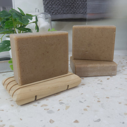 Three soaps are presented on a bathroom countertop.  One soap is to the left and stands on a wooden soap rack.  To the right, another lays flat on the bench, with a final bar standing upright on it.  The soap are all a tan colour with brown flecks.