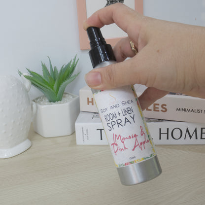A hand holding a room and linen spray bottle with a silver body, black spray top, and a white label reading 'Soy and Shea Room + Linen Spray' with 'Mimosa & Pink Apple' written in pink script. The background shows a light wood tabletop with books, a small potted succulent, and a white ceramic object.