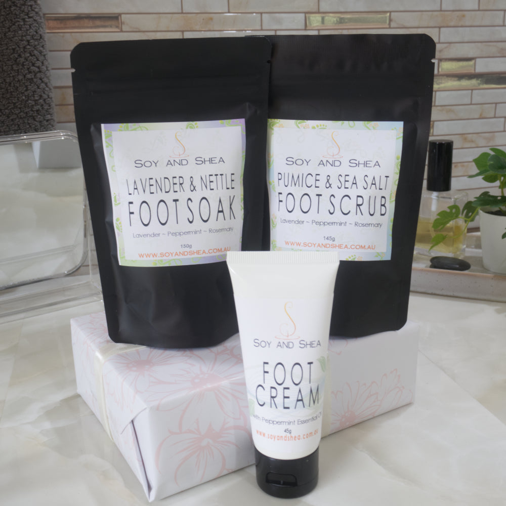 Two black stand up pouches with product labels listing them as Foot Soak and Foot Scrub are standing on box that is wrapped ina white paper with pink floral design.  In front is a white tube of Foot Cream.  Behind is a bathroom counter top