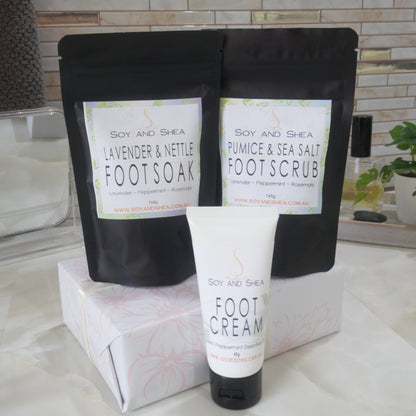 Two black stand up pouches with product labels listing them as Foot Soak and Foot Scrub are standing on box that is wrapped ina white paper with pink floral design.  In front is a white tube of Foot Cream.  Behind is a bathroom counter top