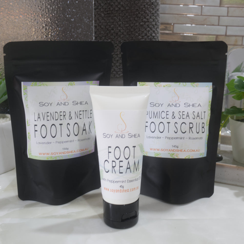 A variety of Foot Care products stand on a marble bathroom countertop with a plant and towel in the background.   The set includes two black standup pouches, one labelled Foot Soak and the other Foot Soak.  In front is a white tube of Foot Cream