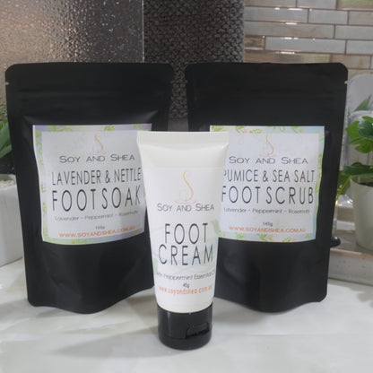 A variety of Foot Care products stand on a marble bathroom countertop with a plant and towel in the background.   The set includes two black standup pouches, one labelled Foot Soak and the other Foot Soak.  In front is a white tube of Foot Cream