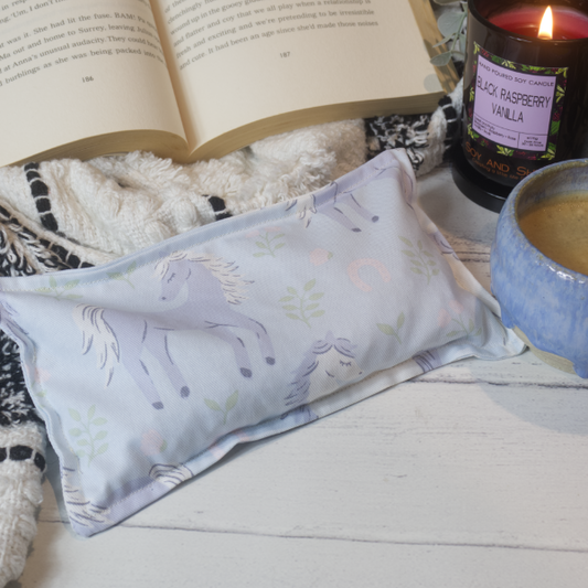 A mini rectanglula heat pack with a pale blue bacground and illustrations of horse dark blue and white horses, placed on a wooden surface next to an open book and a lit candle.