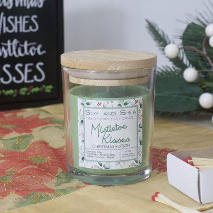 A clear candle jar with wooden lid sits on a sideboard surrounded by a Christmas sign and wreath.  The jar is filled with green wax and features a decorative product label announcing this has Mistletoe Kisses