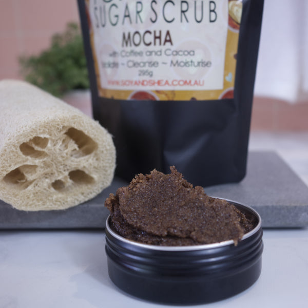 A zoomed in image of a black tin filled with brown sugar scrub sitting on a white bench with the blurred image of the packaging in the background.