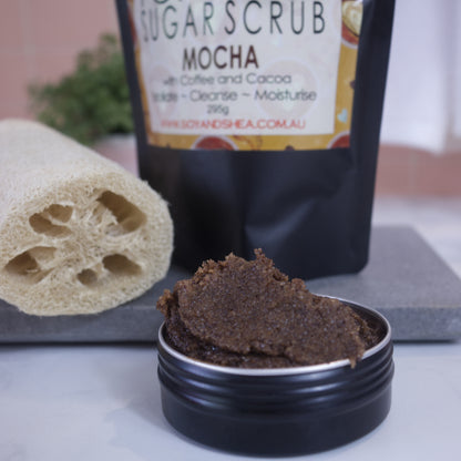 A zoomed in image of a black tin filled with brown sugar scrub sitting on a white bench with the blurred image of the packaging in the background.