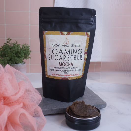 A black zip lock pouch stands up-right on a grey stone tray in front of pink tiled wall.  The Label reads  FOAMING SUGAR SCRUB MOCHA against a coffee themed background.    In front is a small black tin overflowing with a brown scrub.