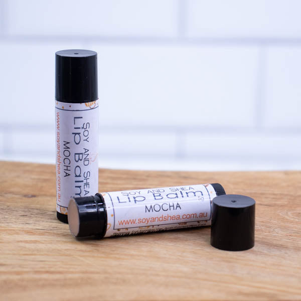 Two black tubes of lip balm are displayed on a wooden surface with blurred tiled background.  One tube stands on its end while the other lays flat in front with the cap removed revealing a tan balm.  The label is white with a random pattern of coffee cups.